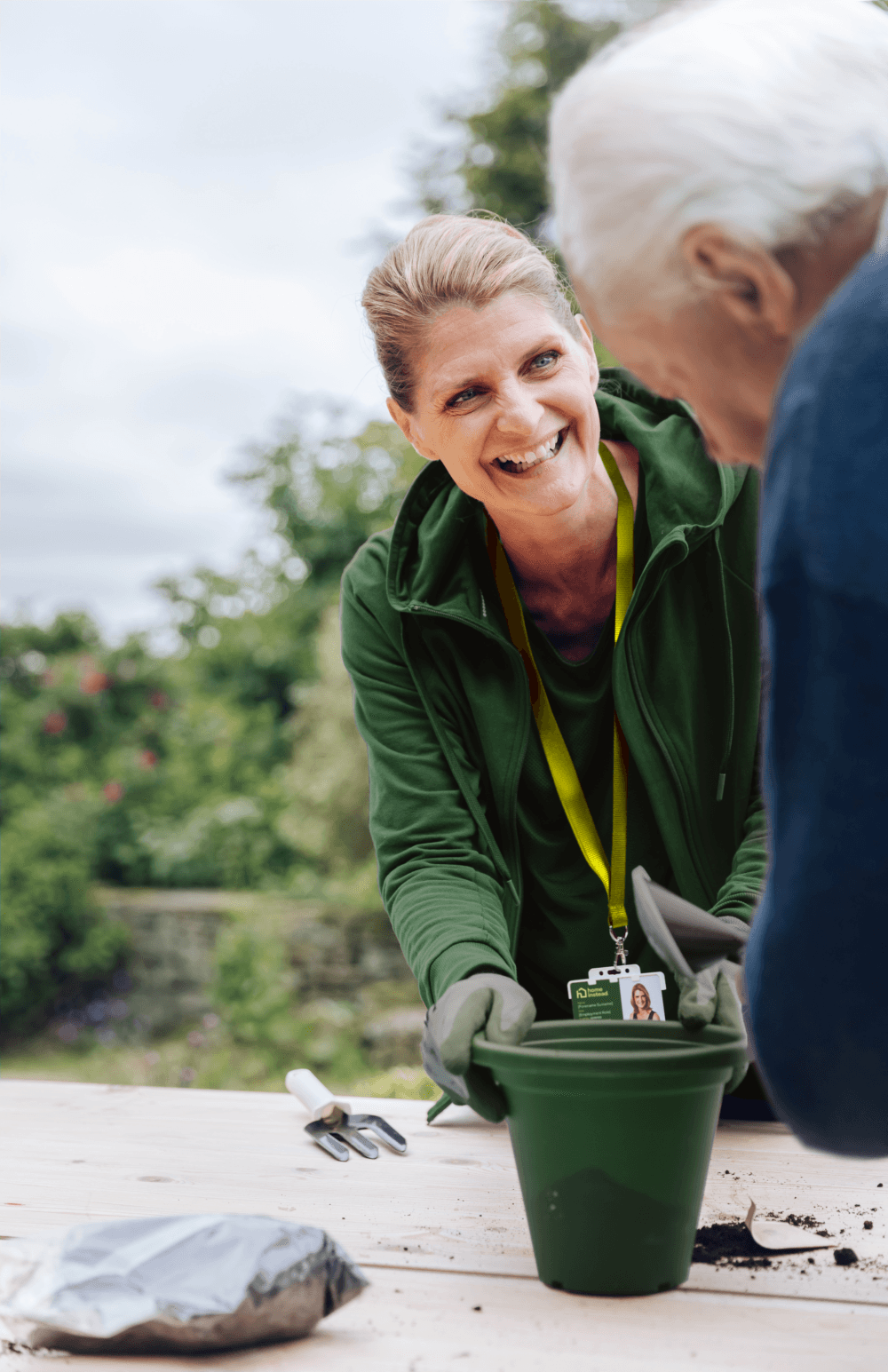 Smiling woman helps an older adult with planting in a garden, holding a green pot. - Home Instead