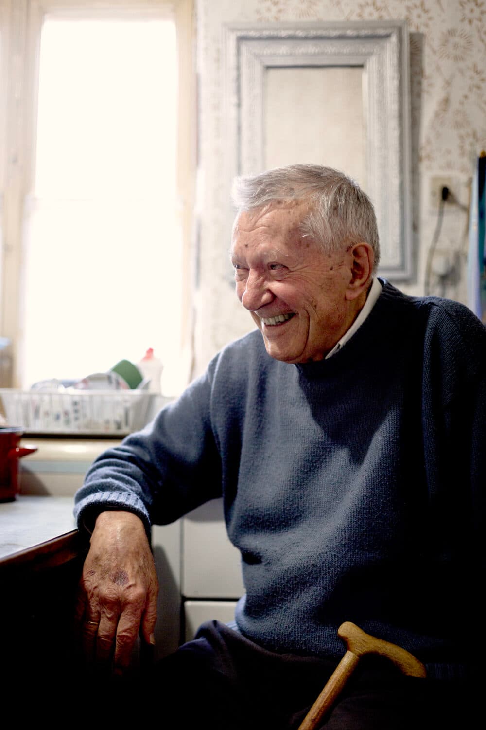 An elderly man with a cane smiles while sitting in a sunlit room with framed art behind him. - Home Instead