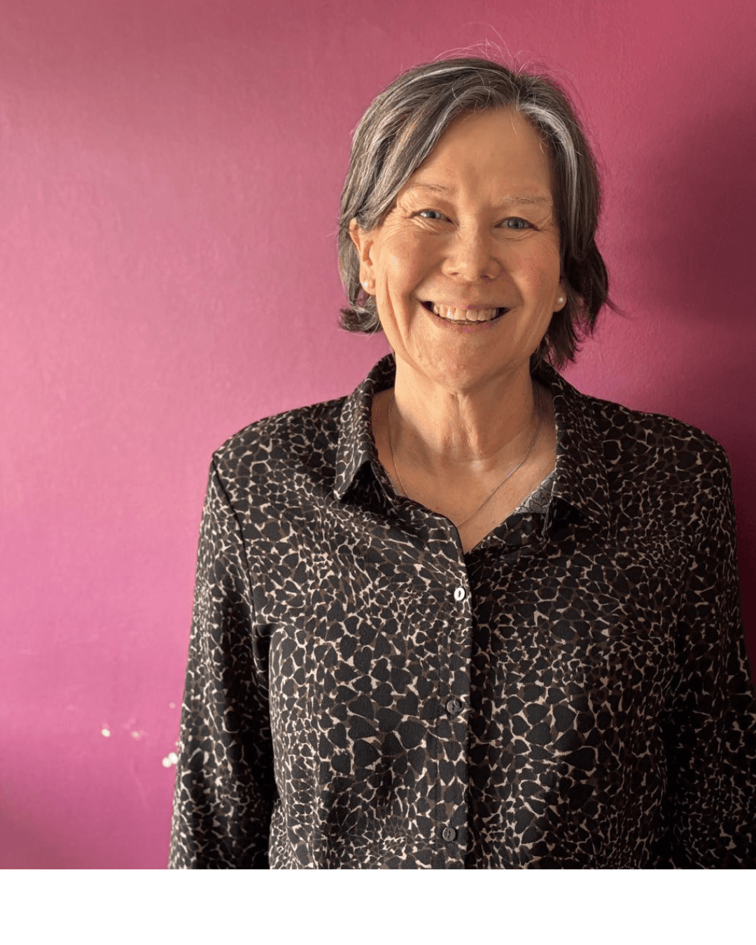 Smiling woman with short gray hair in a patterned shirt stands in front of a pink wall. - Home Instead