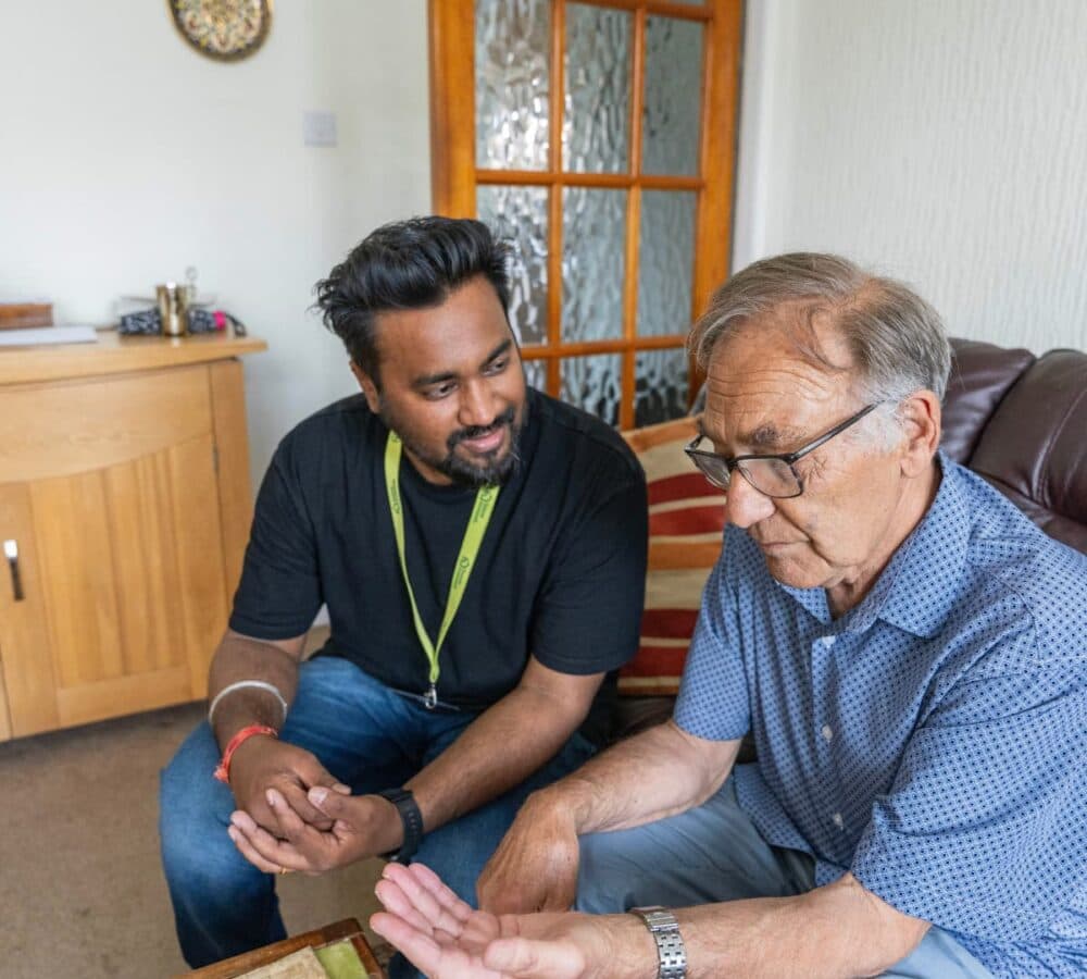 An older male adult wearing eyeglasses and with grey hair and sitting on a couch inisde the house chatting with his younger male carer with black hair and mustache