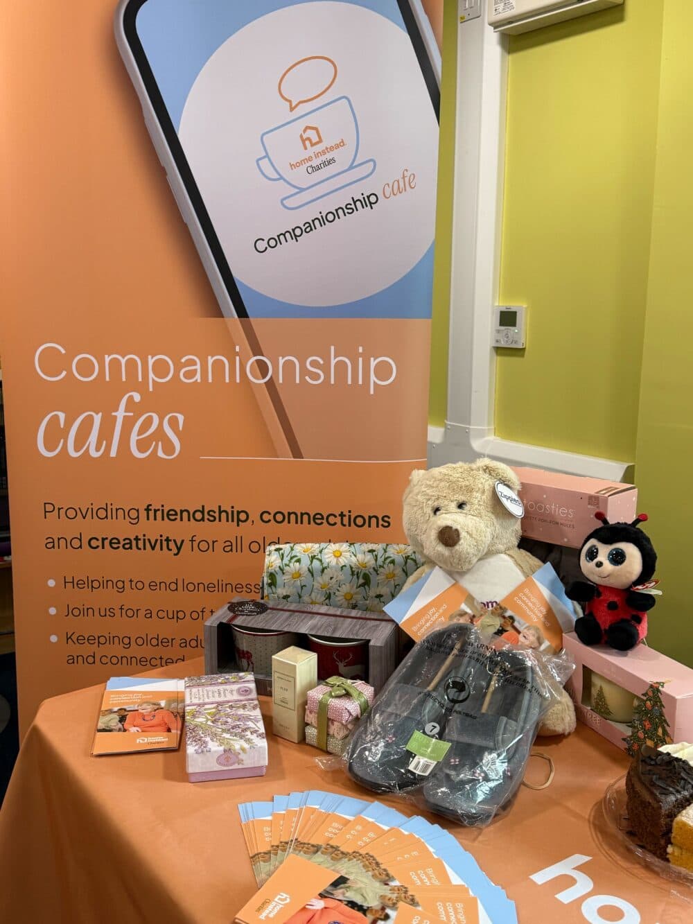 A display table with gifts and brochures in front of a "Companionship cafés" sign promoting friendship. - Home Instead