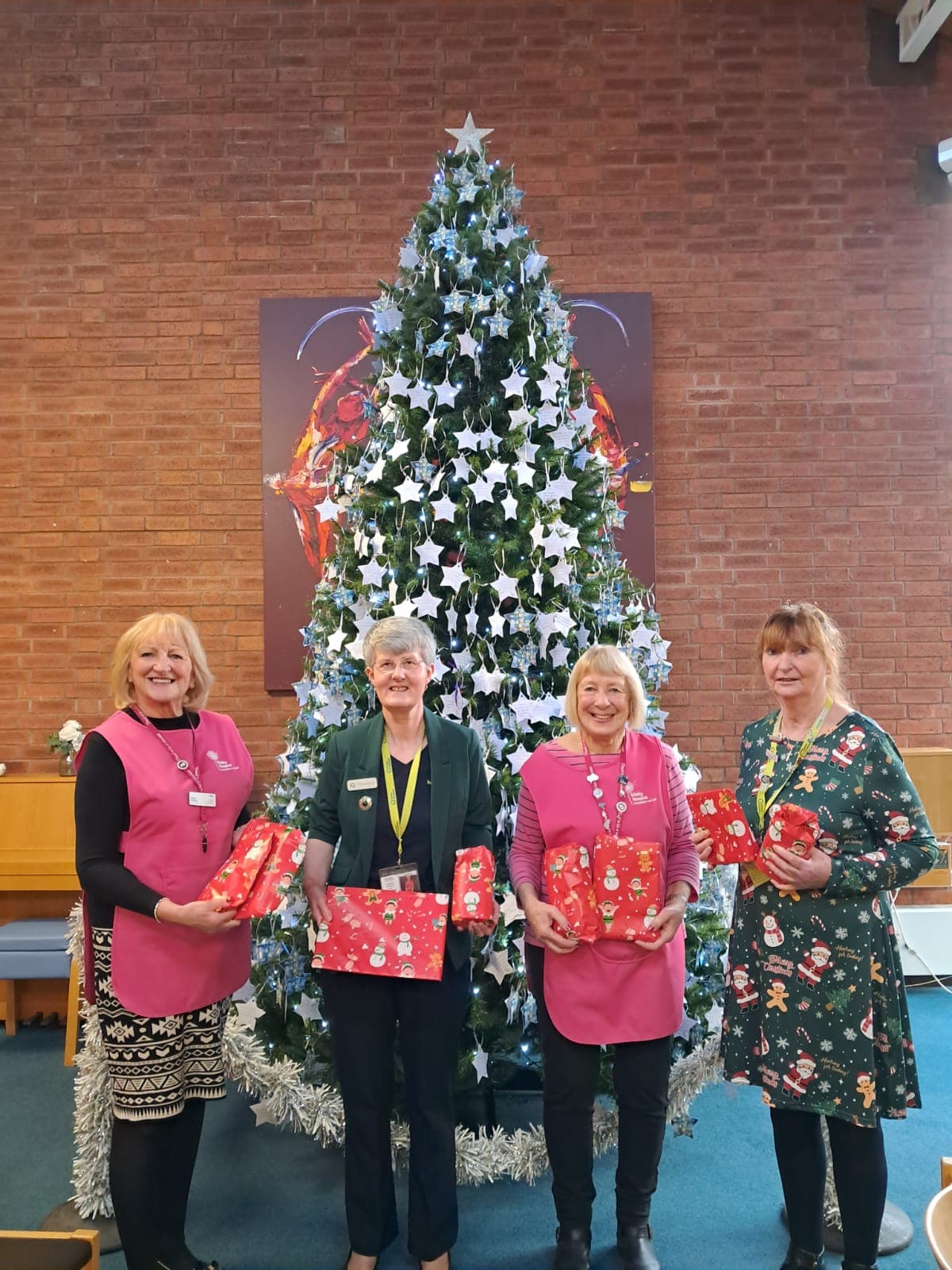 Four women standing in front of a decorated Christmas tree, holding wrapped presents and smiling. - Home Instead