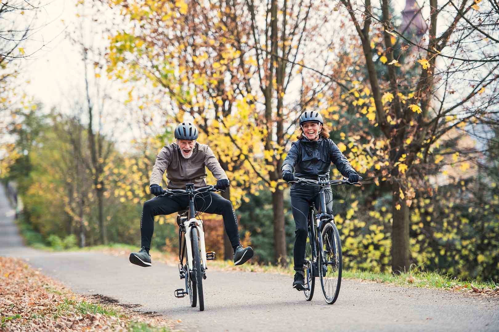 Two people riding bicycles on a tree-lined path in autumn, both smiling and wearing helmets. - Home Instead