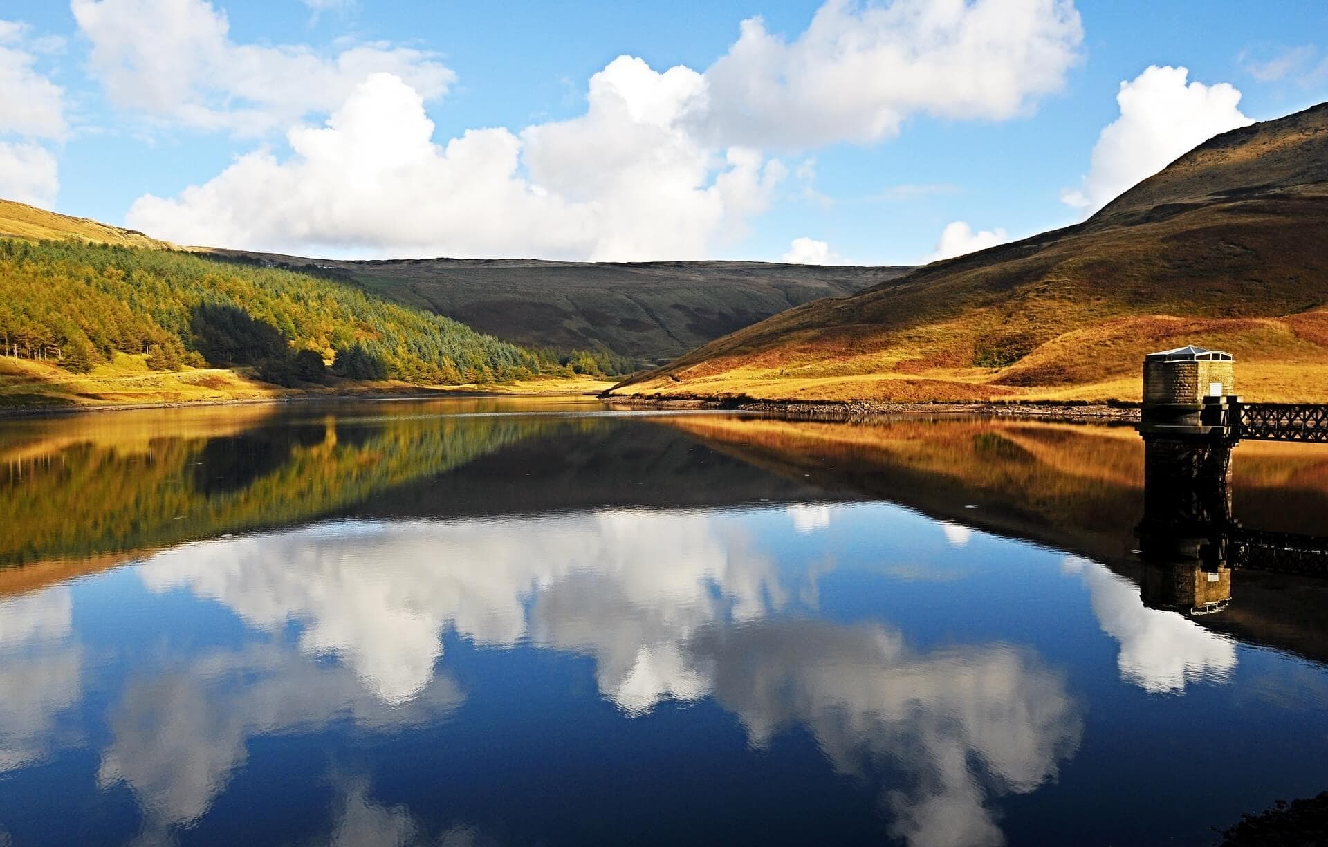 A serene lake with reflections of clouds and hills; a small hut is on the shore under a bright blue sky. - Home Instead