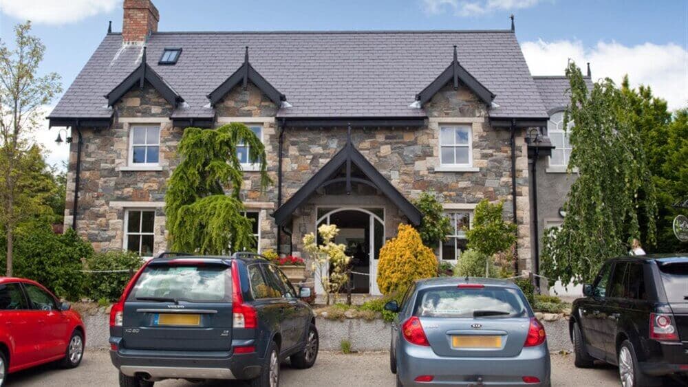 A stone house with a gray roof, surrounded by plants and trees, and several cars parked in front of it. - Home Instead