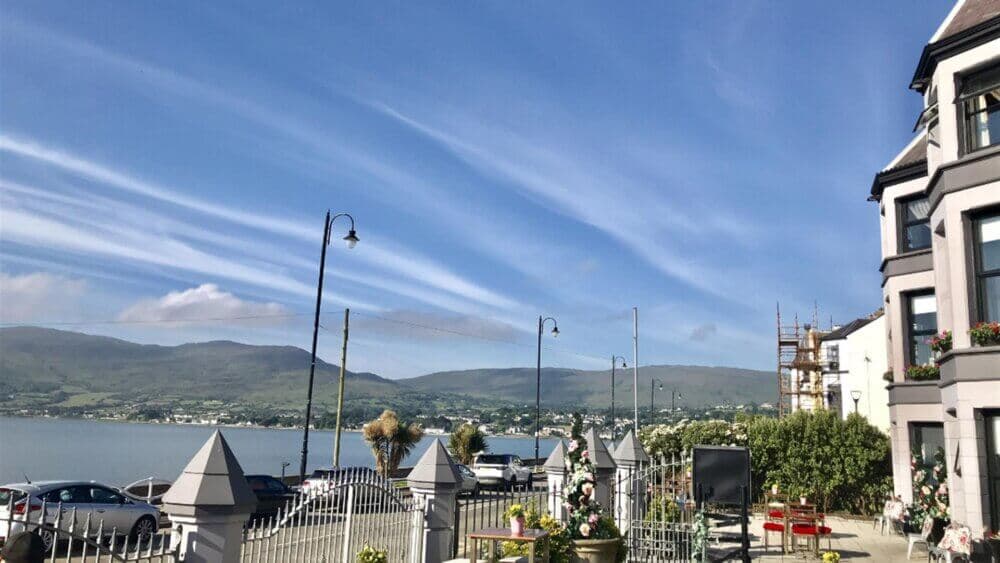 A scenic view of a waterfront town with mountains in the background, houses, parked cars, and a clear blue sky. - Home Instead