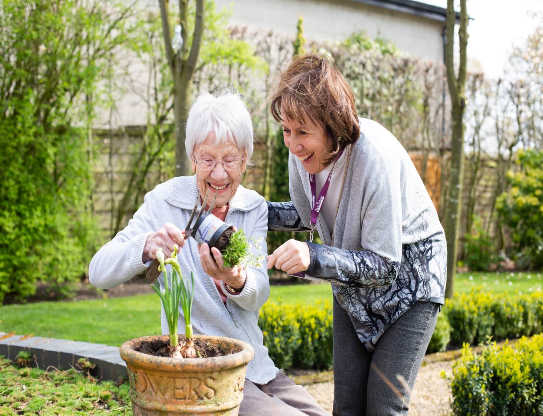 Two women gardening together, one elderly and one younger, both smiling and enjoying planting flowers outside. - Home Instead