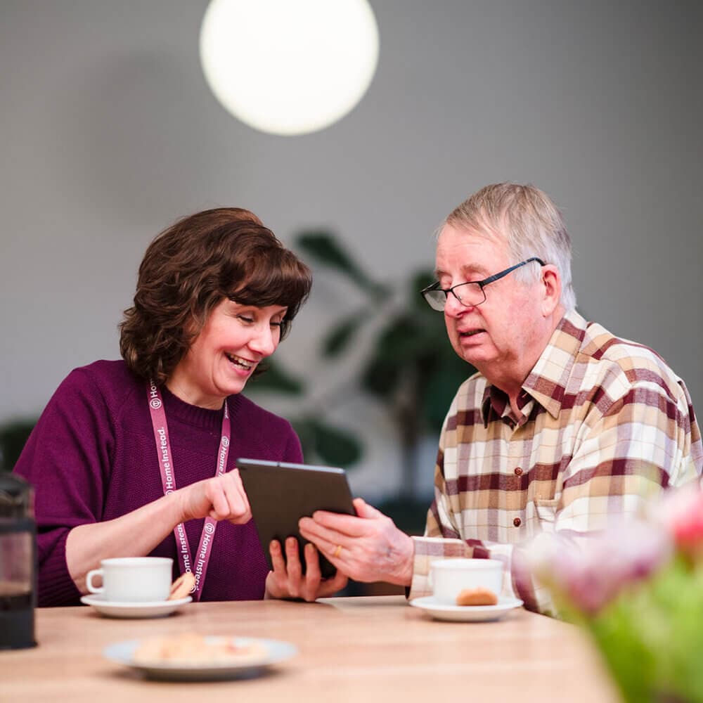 A woman shows an older man something on a tablet while they sit at a table with coffee cups. - Home Instead