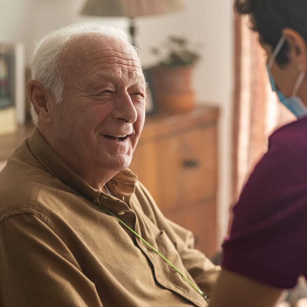An elderly man smiles as he talks with a caregiver wearing a face mask in a warmly lit room. - Home Instead
