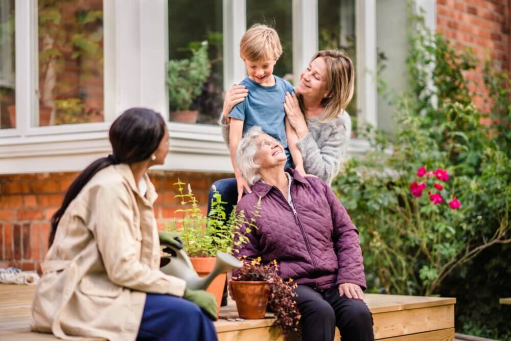 A young boy and three women share a moment on a garden patio, surrounded by plants and flowers. - Home Instead