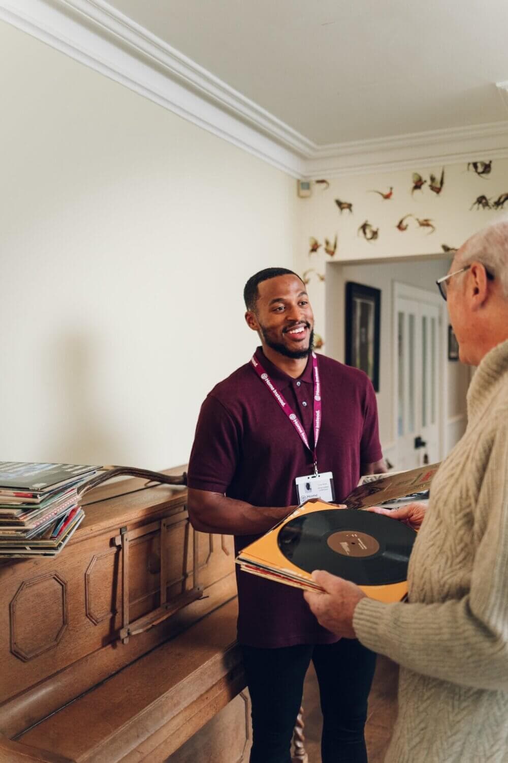 A man in a maroon shirt hands a vinyl record to an elderly man by a wooden piano with more records on top. - Home Instead