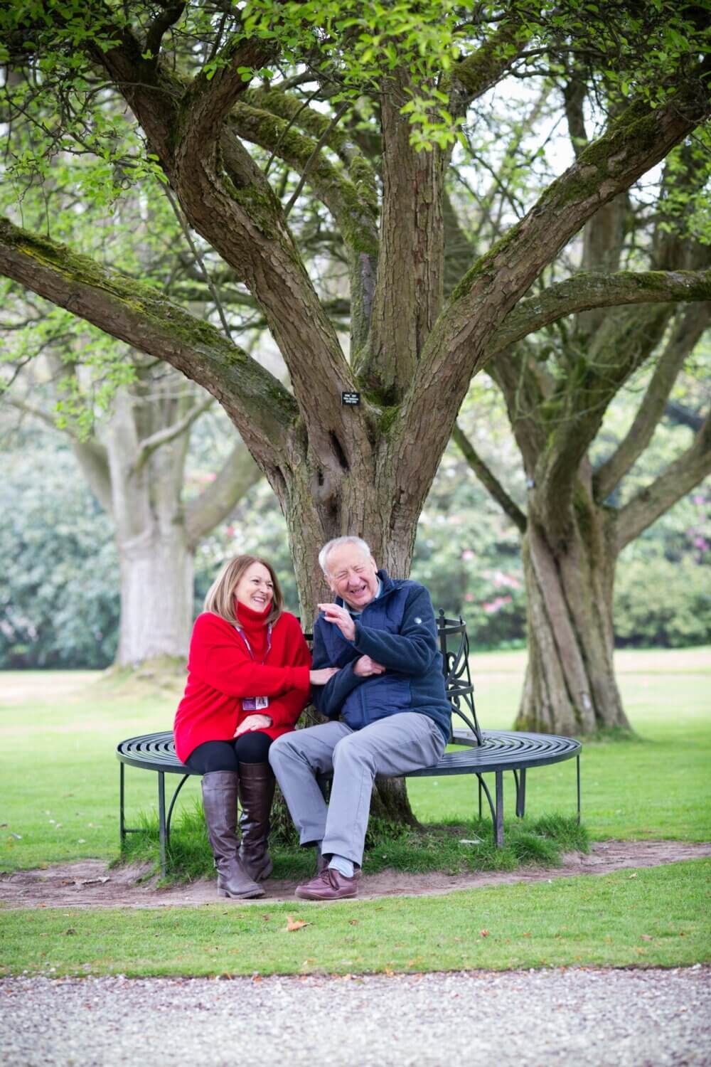 An elderly couple sitting on a wrap-around bench under a large tree, laughing and enjoying each other's company. - Home Instead