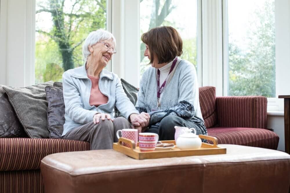 Two women smiling and holding hands on a couch. A wooden tray with tea set is on an ottoman in front of them. - Home Instead