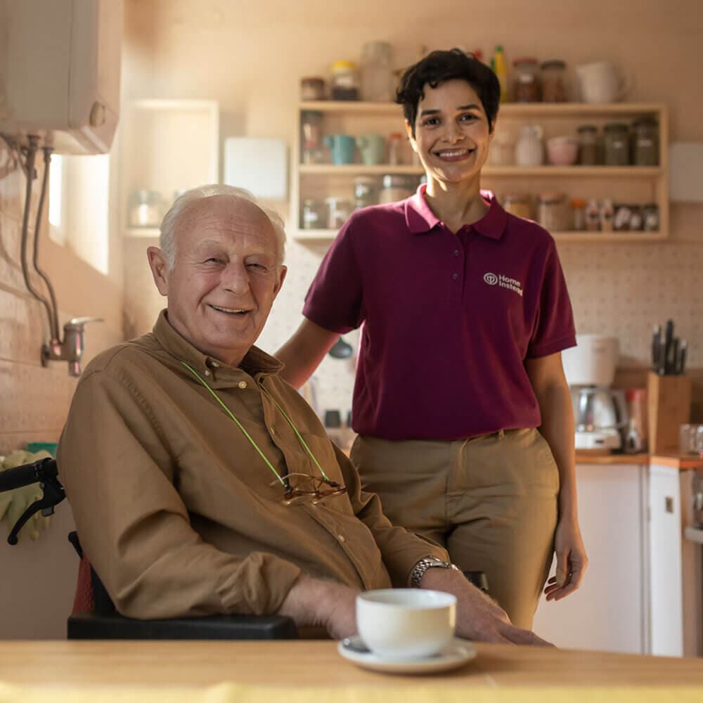 A smiling elderly man sits at a table with a young woman in a uniform standing beside him in a bright kitchen. - Home Instead