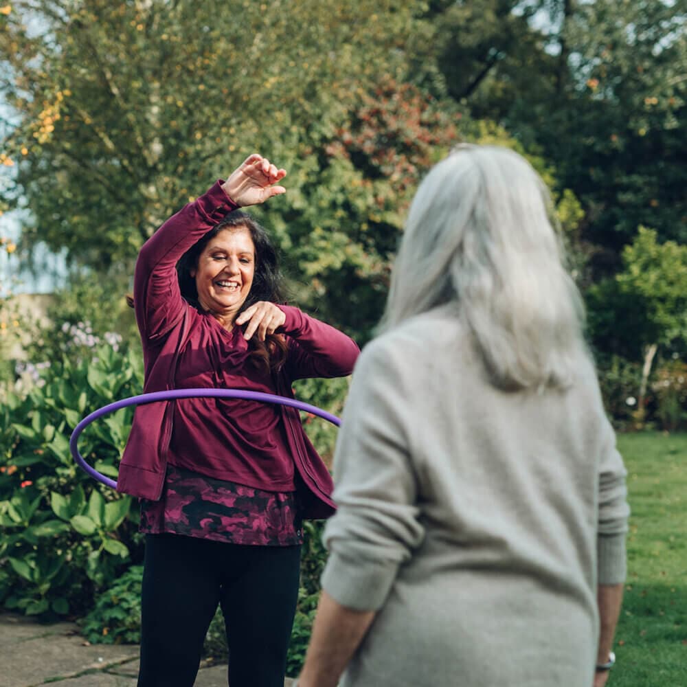A smiling woman hula hoops outside as another older woman with gray hair looks on in a garden. - Home Instead