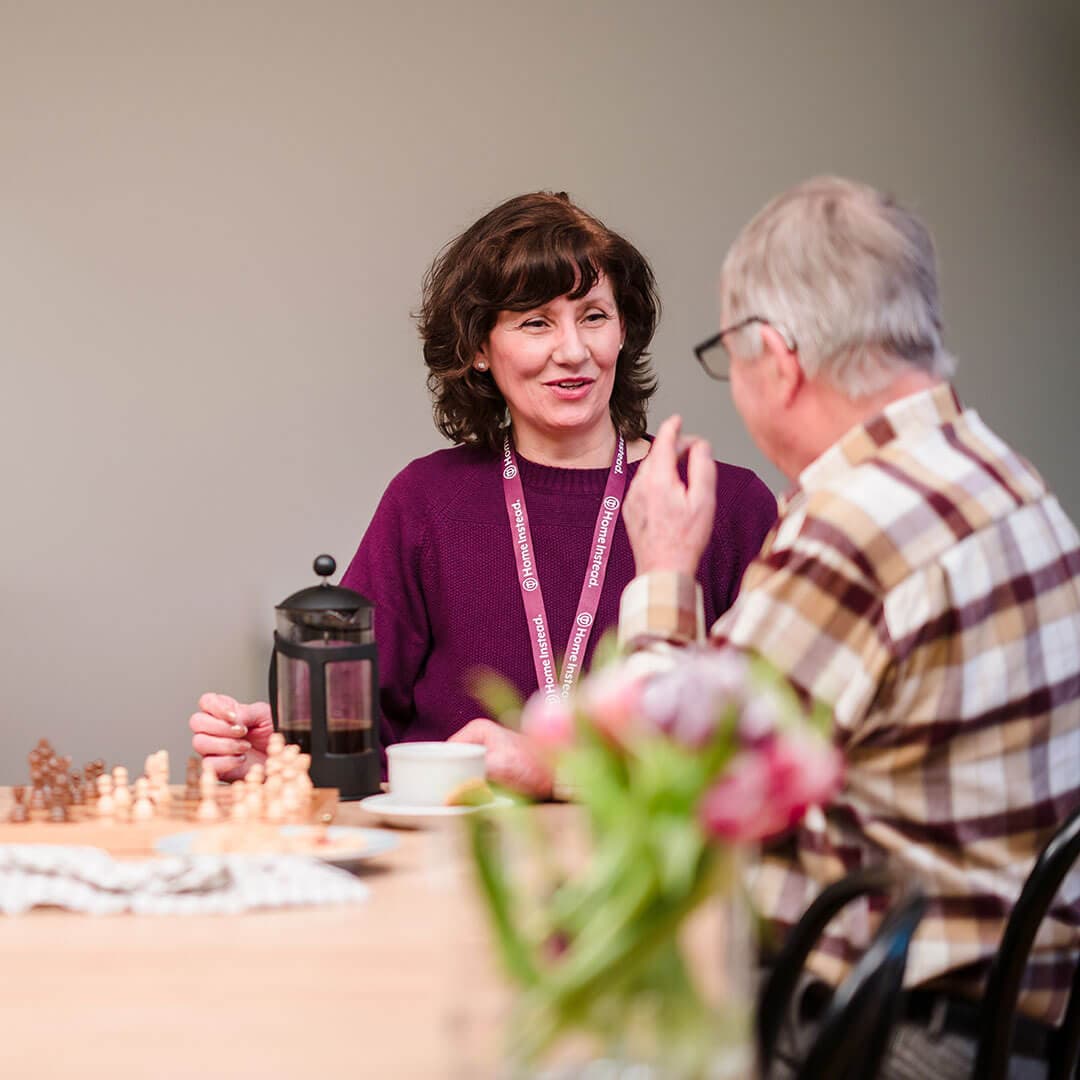 Woman and elderly man having a conversation at a table with coffee and chess pieces. - Home Instead