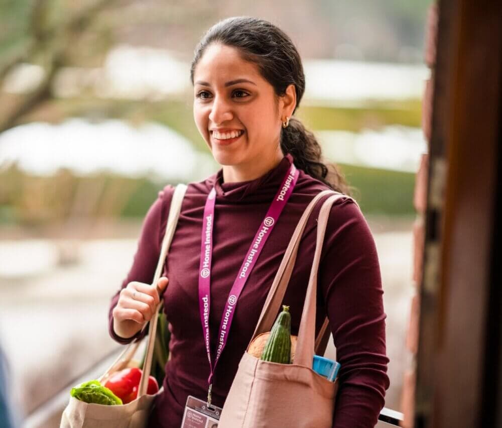 A smiling woman carrying grocery bags stands at a door, wearing a lanyard with ID and a dark red sweater. - Home Instead
