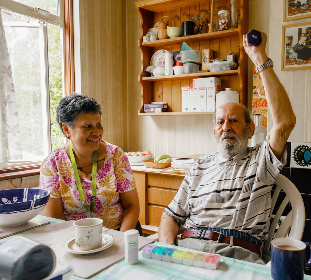 An older male adult with grey hair exercising while sitting on a chair inside the kitchen with his younger female carer with short hair happy and smiling - Home Instead