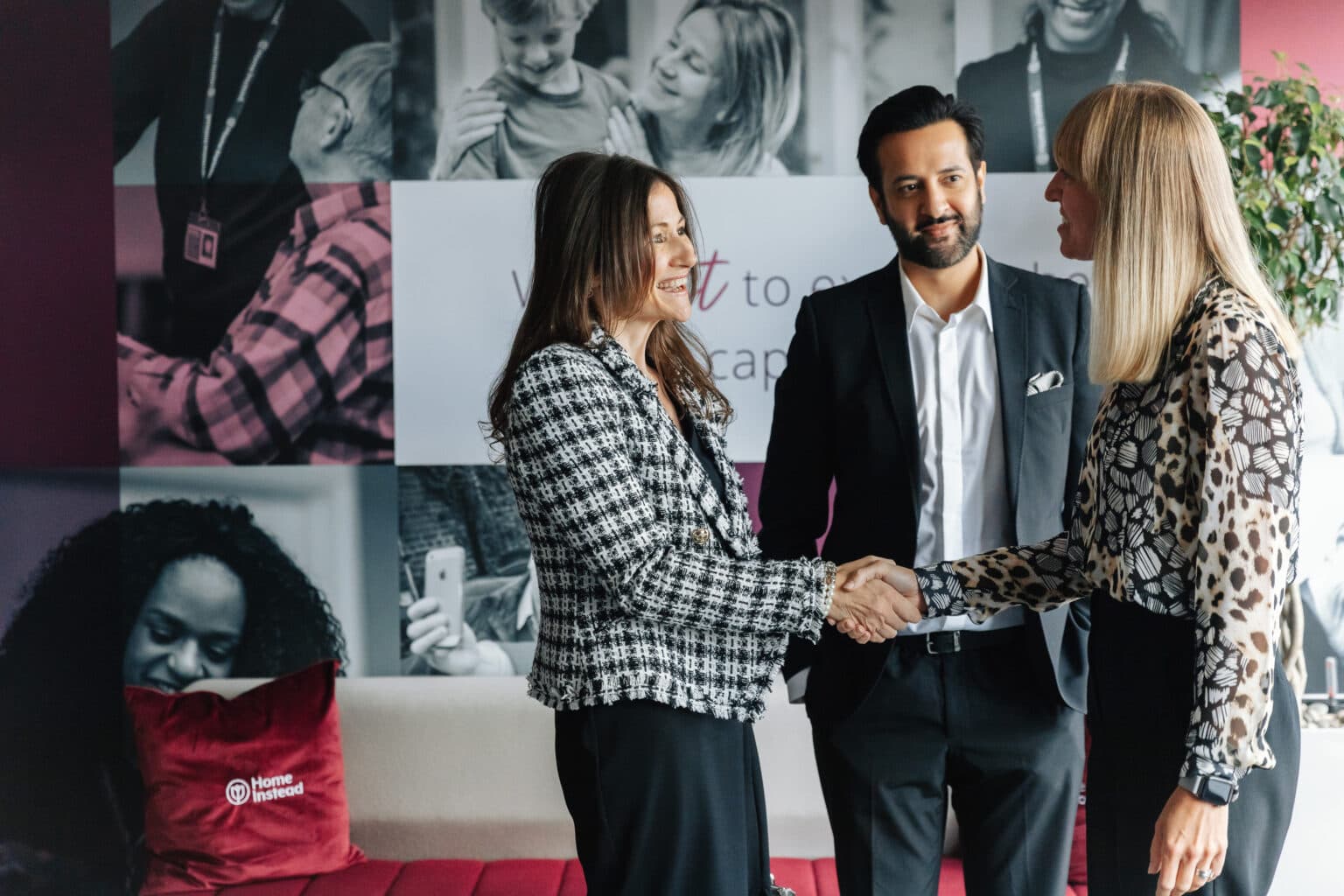 Three people in business attire greet each other with a handshake in a modern office with photo wall décor. - Home Instead