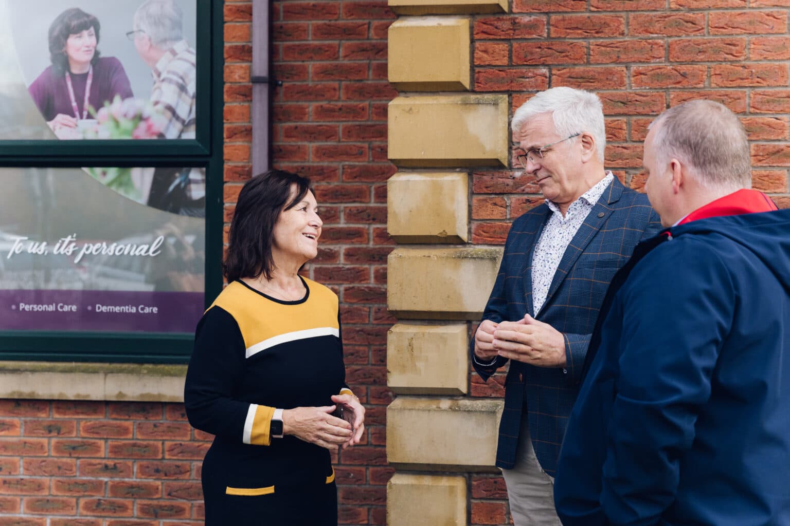 Three adults chatting outside a brick building near a care home sign, smiling and engaged in conversation. - Home Instead