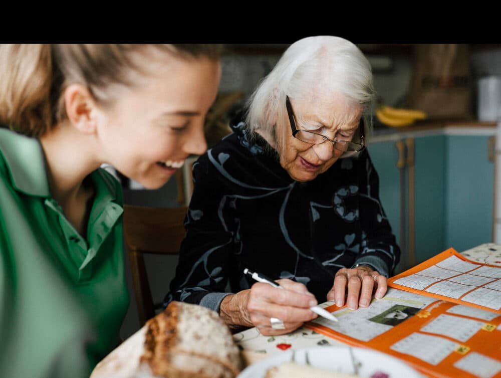 A young woman smiles as she watches an elderly woman fill in a diary with a pen. - Home Instead