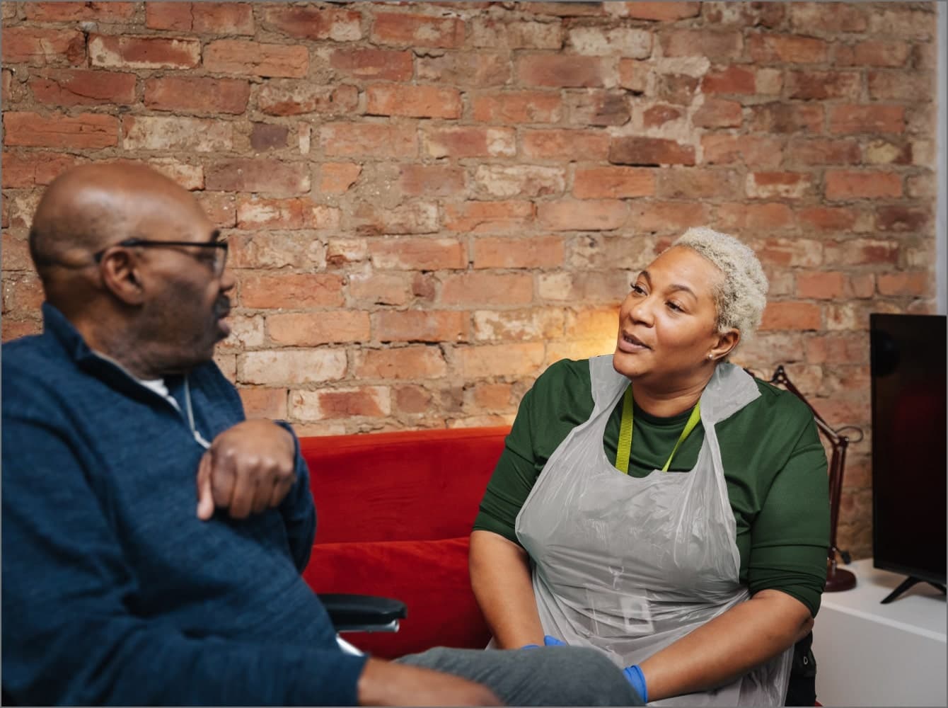 A smiling woman with a name badge talks to an older person sitting across from her in a bright room with plants. - Home Instead