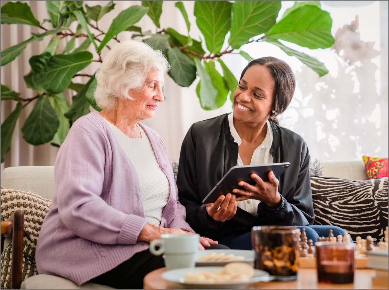 An elderly woman and a younger woman sit together on a couch, smiling and looking at a smartphone. - Home Instead