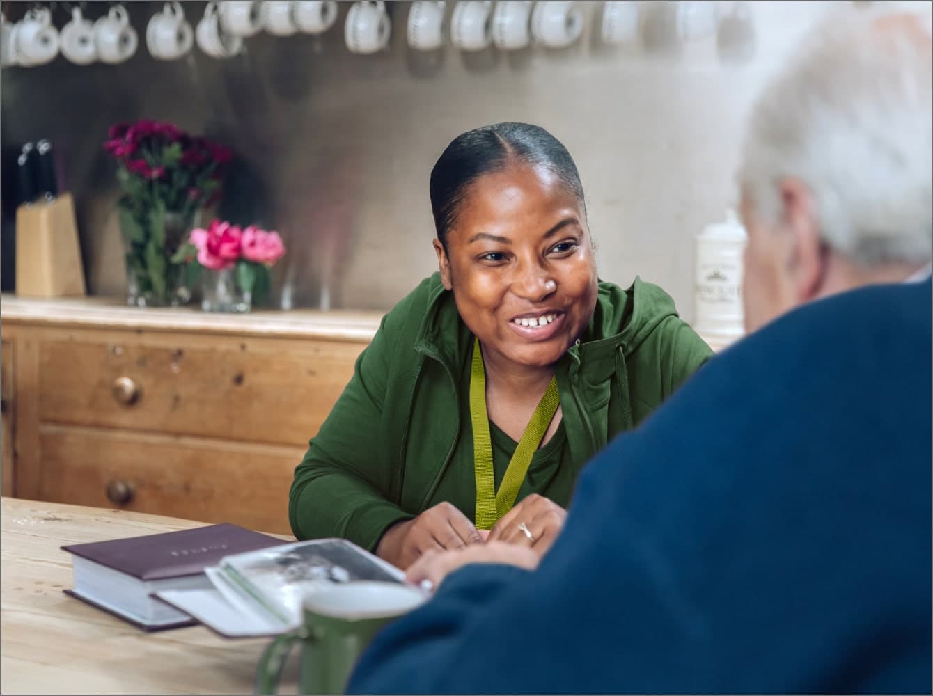 An elderly woman smiles while sitting at a wooden table in a café, with a cup of coffee in front of her. - Home Instead