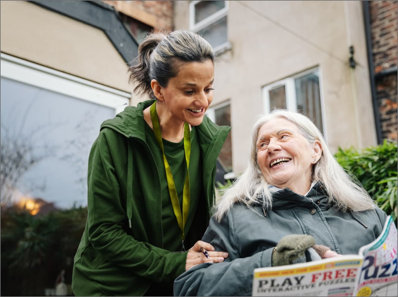 An elderly woman smiles while holding a plant as she talks to a seated younger person in an outdoor garden. - Home Instead