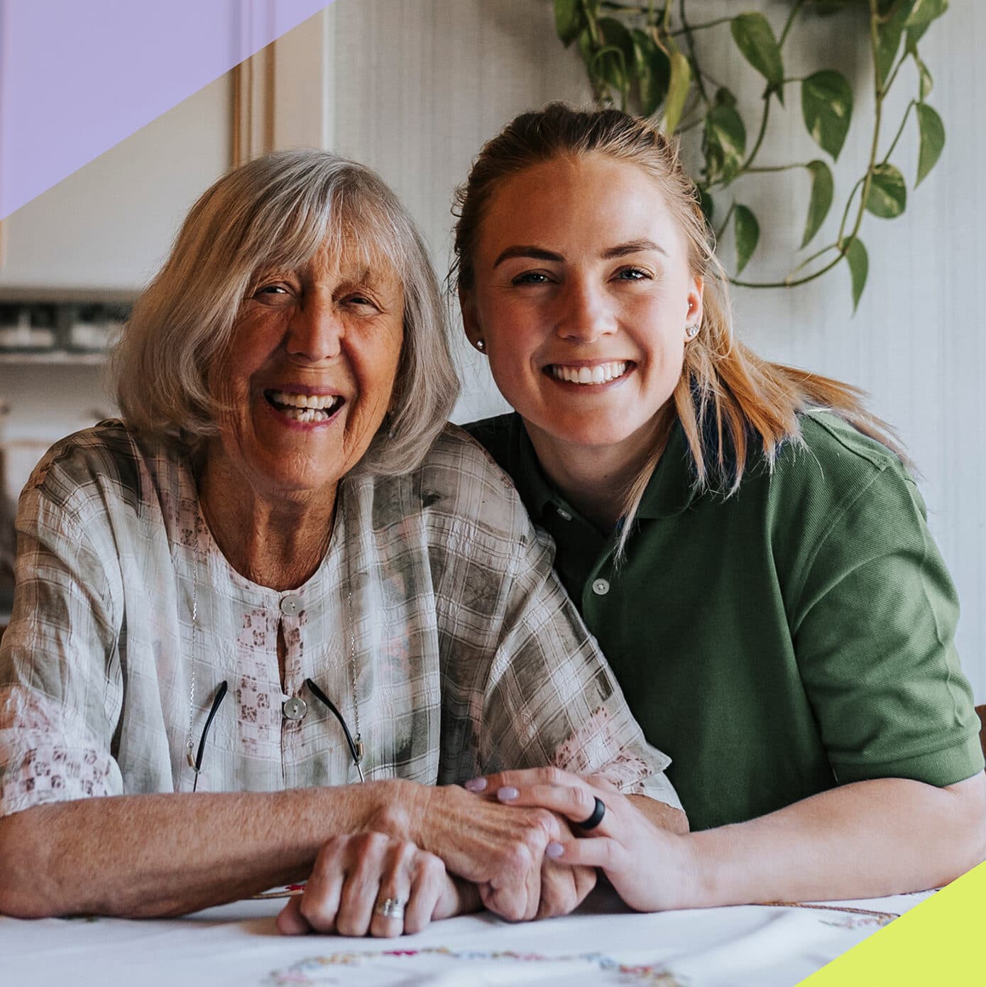 An elderly woman and a young woman smiling together at a table, holding hands, with a plant in the background. - Home Instead