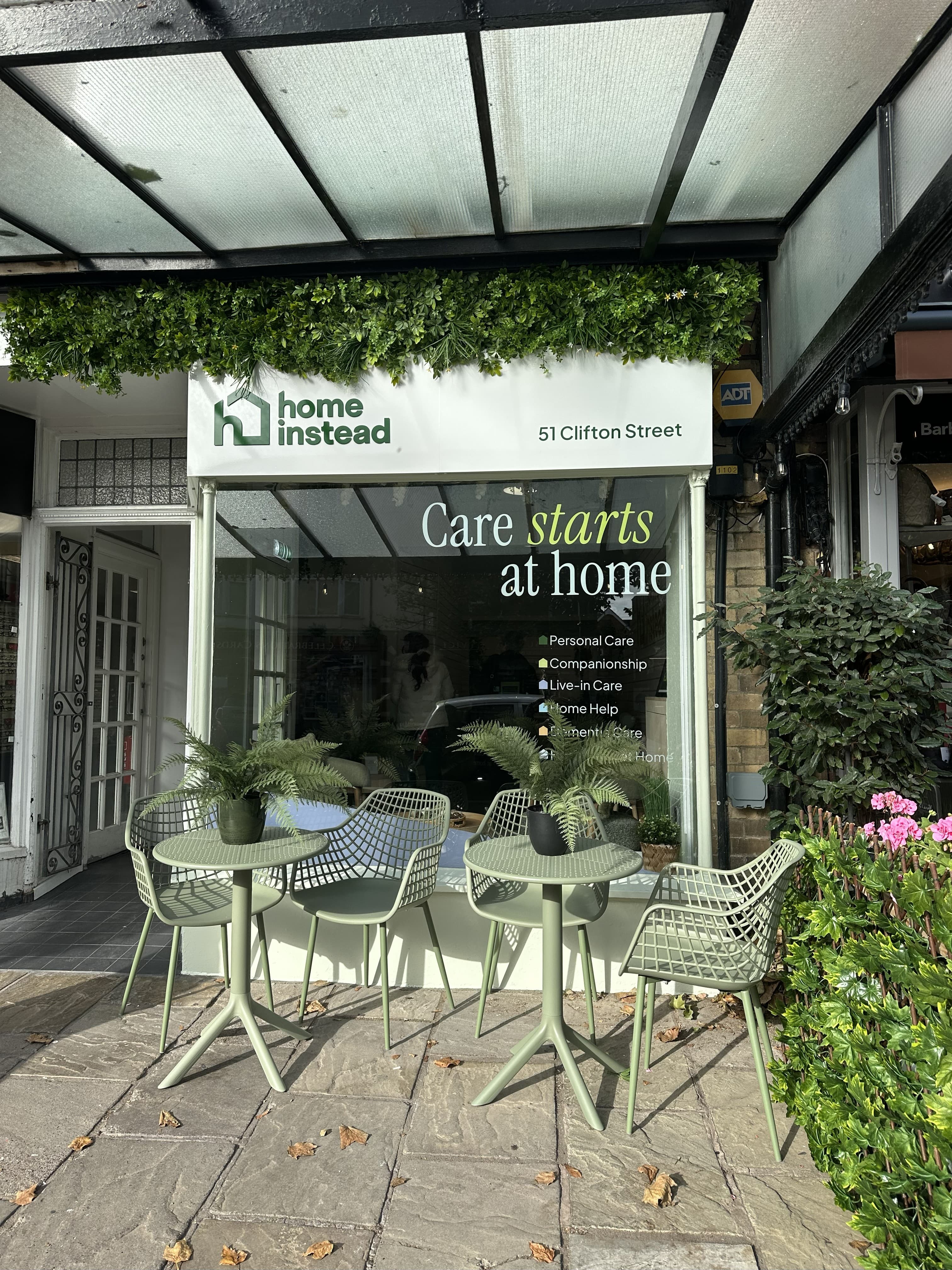 Street view of "Home Instead" care service with green chairs and tables outside under a leafy awning. - Home Instead