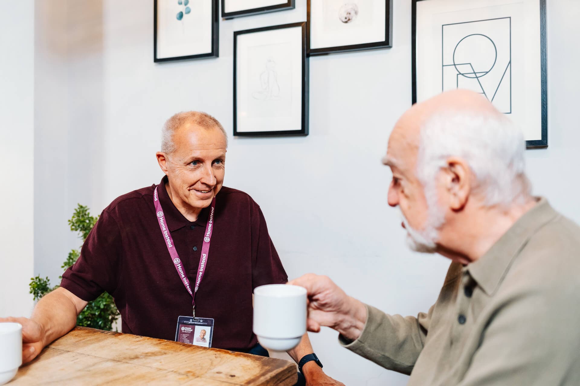 Two elderly men having a conversation while holding coffee cups at a table, with framed pictures on the wall behind them. - Home Instead
