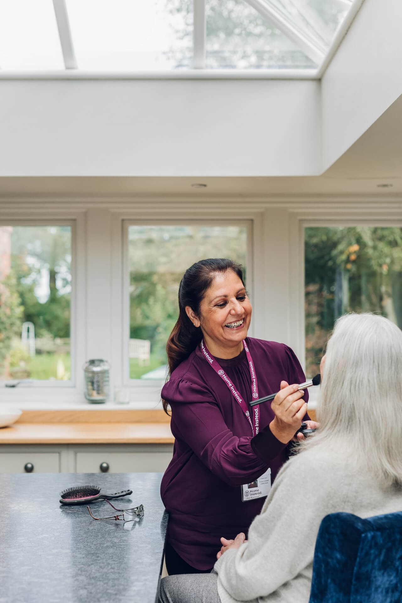 A caregiver in a purple shirt smiles while applying makeup to an elderly woman in a bright, modern kitchen. - Home Instead