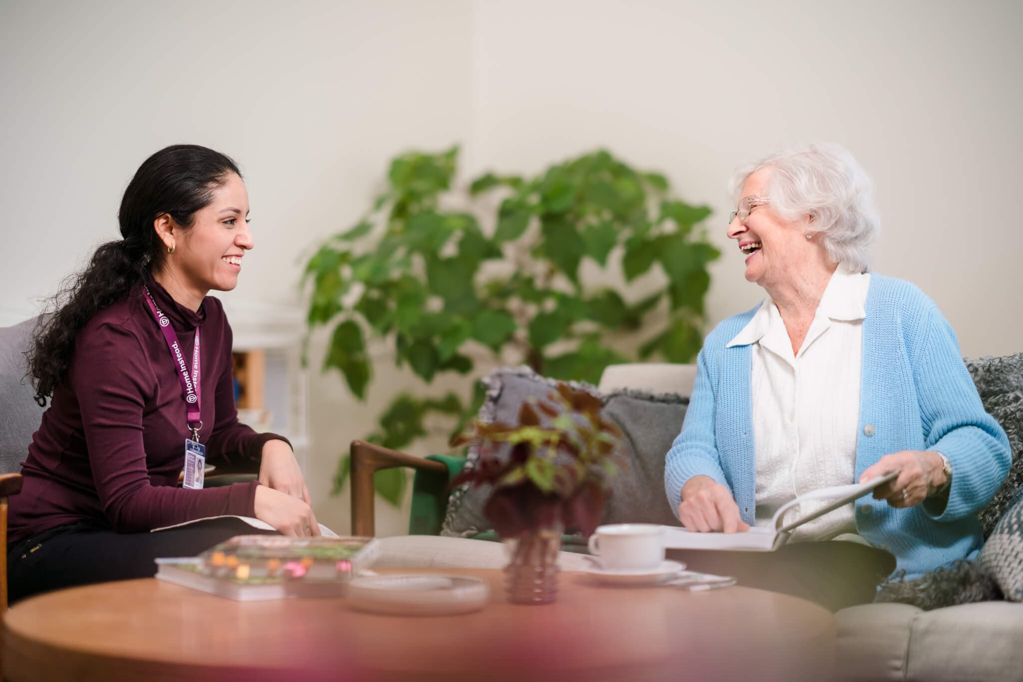 A young woman and an elderly woman smile at each other while sitting and chatting in a cozy living room. - Home Instead