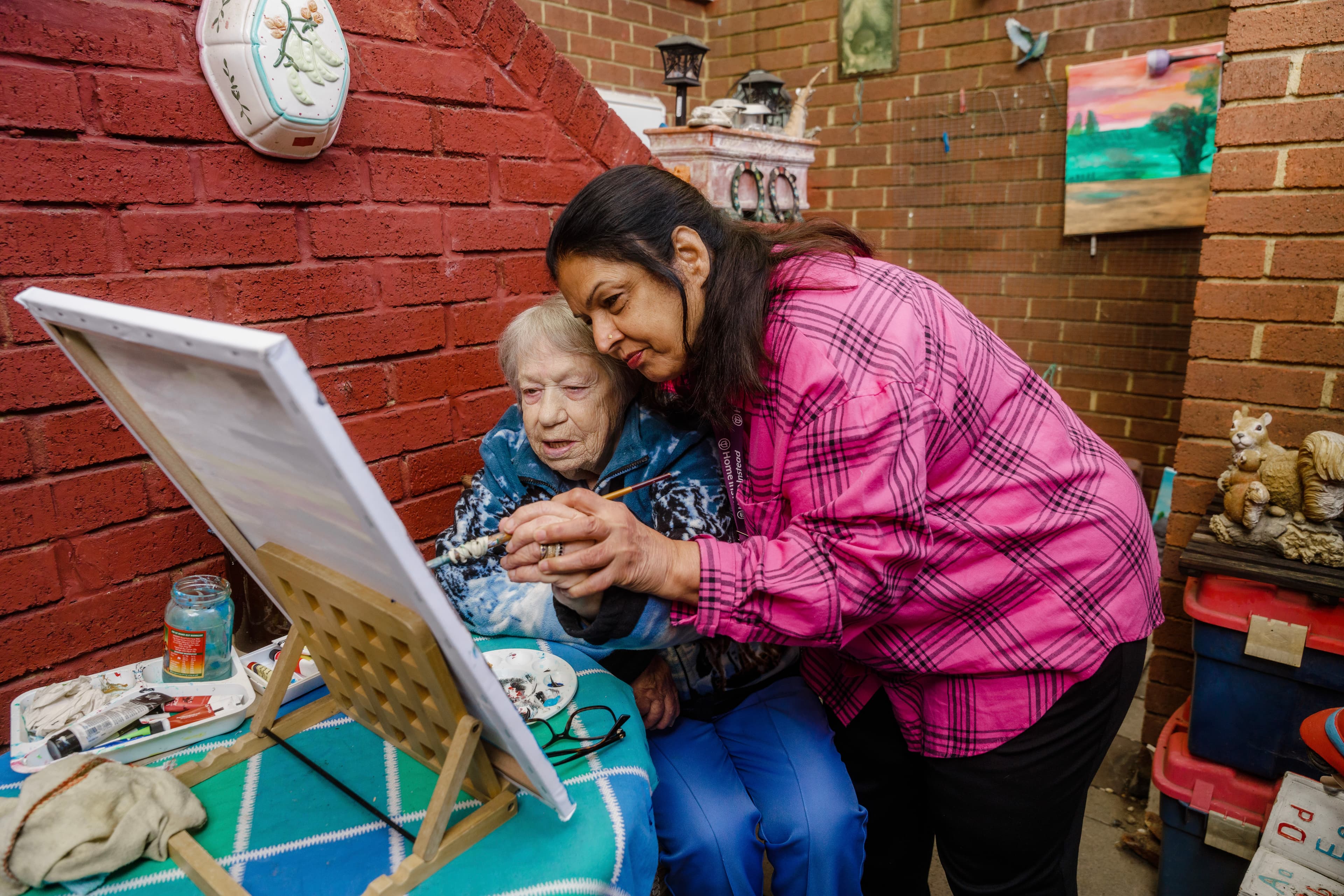 A woman assists an elderly lady at an easel, guiding her hand as they paint together in a brick-walled room. - Home Instead