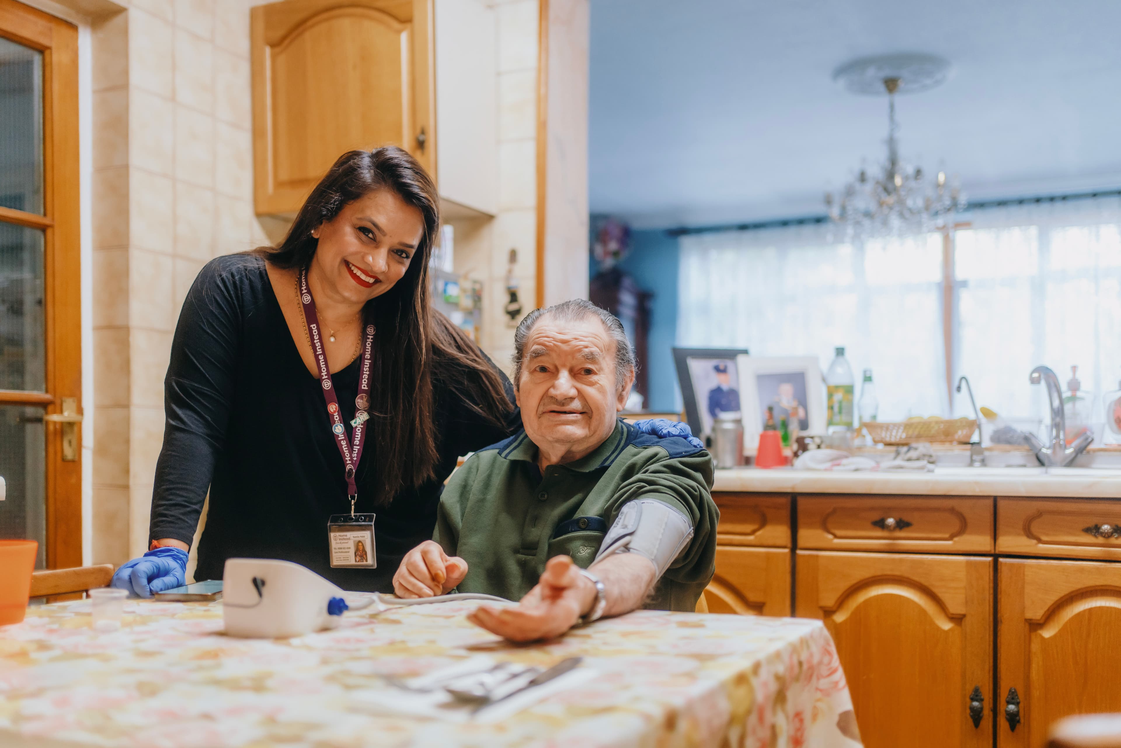 A woman and an elderly man at a kitchen table, with the woman smiling and the man having his blood pressure checked. - Home Instead