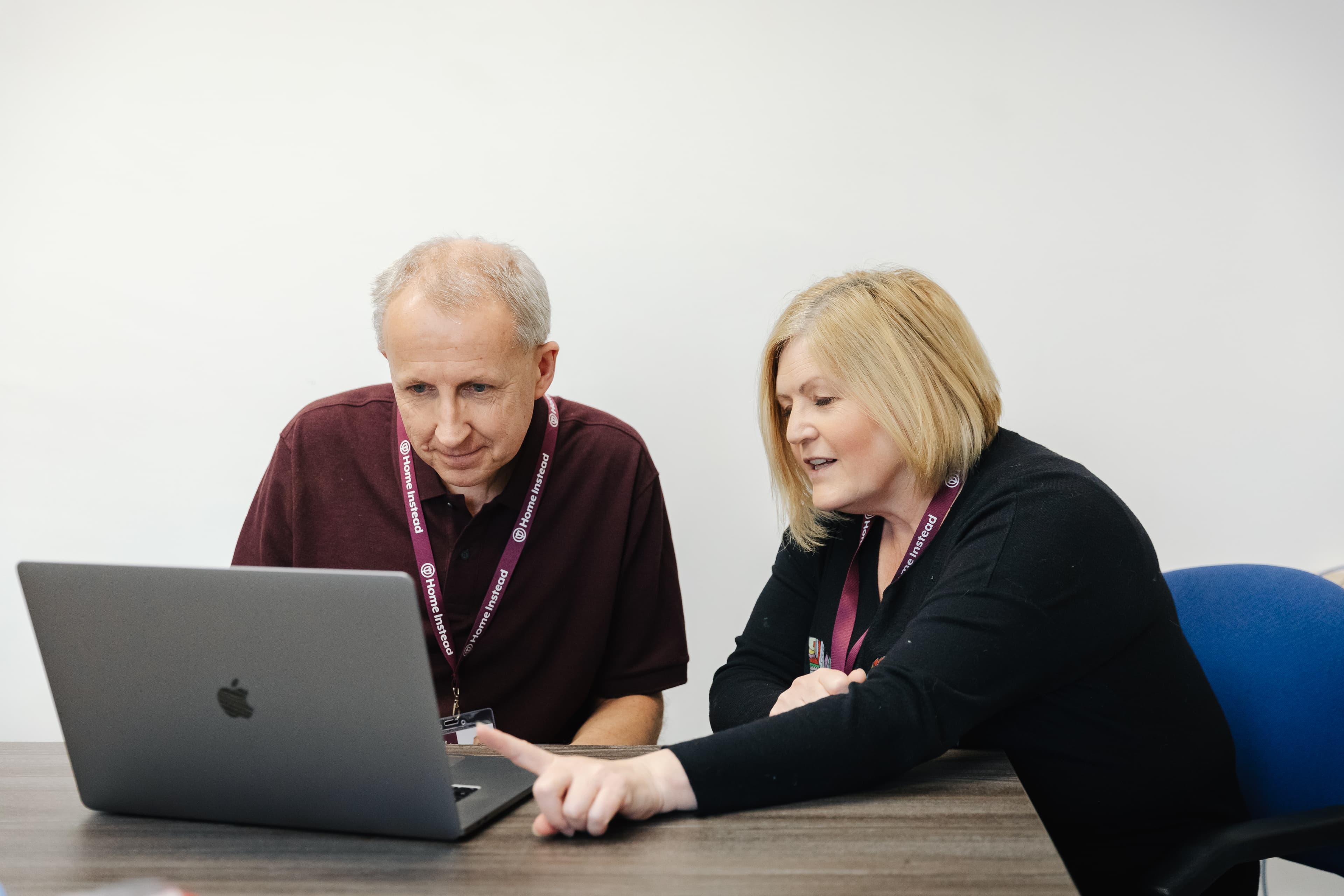 Two people sitting at a desk, looking at a laptop screen, with lanyards around their necks. - Home Instead