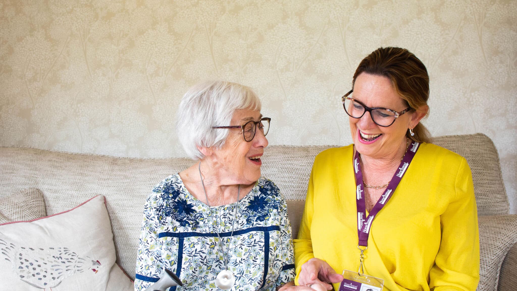 An elderly woman and a caregiver sit on a couch, smiling and chatting together inside a cozy home. - Home Instead