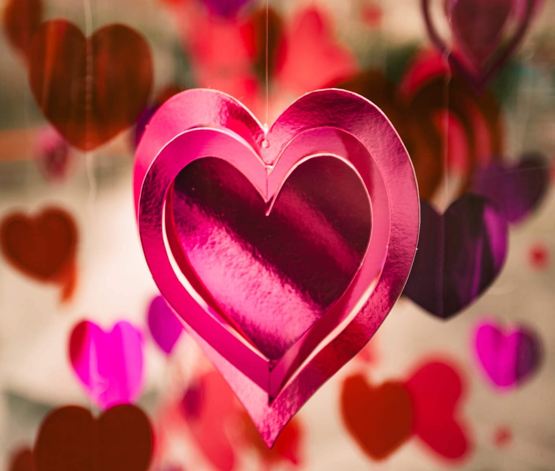 Hanging shiny pink hearts with a blurred background of red and purple hearts in various sizes. - Home Instead
