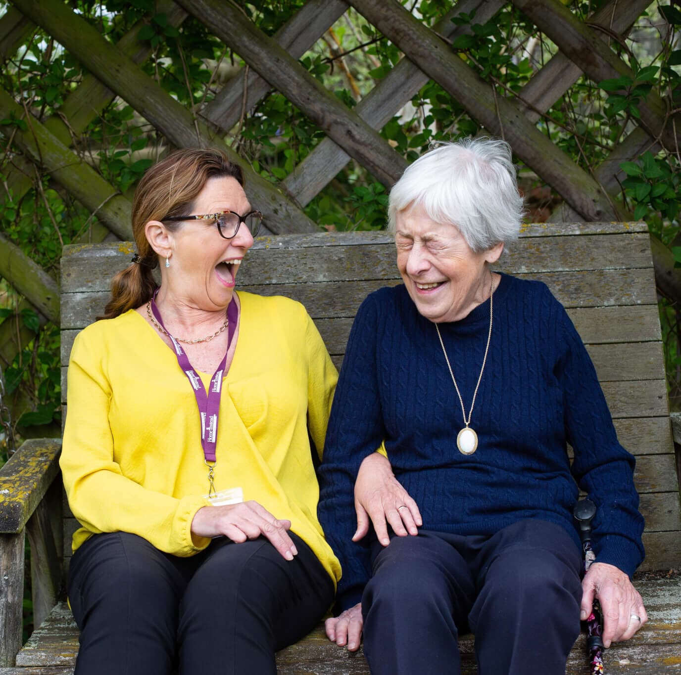 Two women sitting on a wooden bench, laughing and enjoying a moment together against a leafy background. - Home Instead