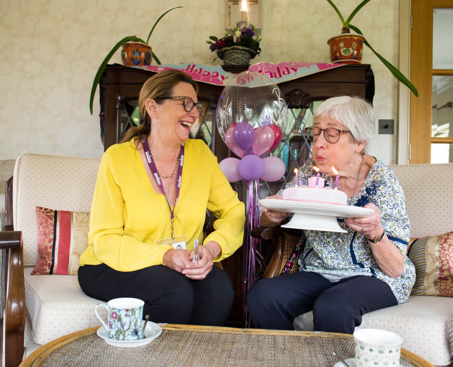 Home Instead client blowing out candles on her birthday cake, watched by her smiling Home Instead carer