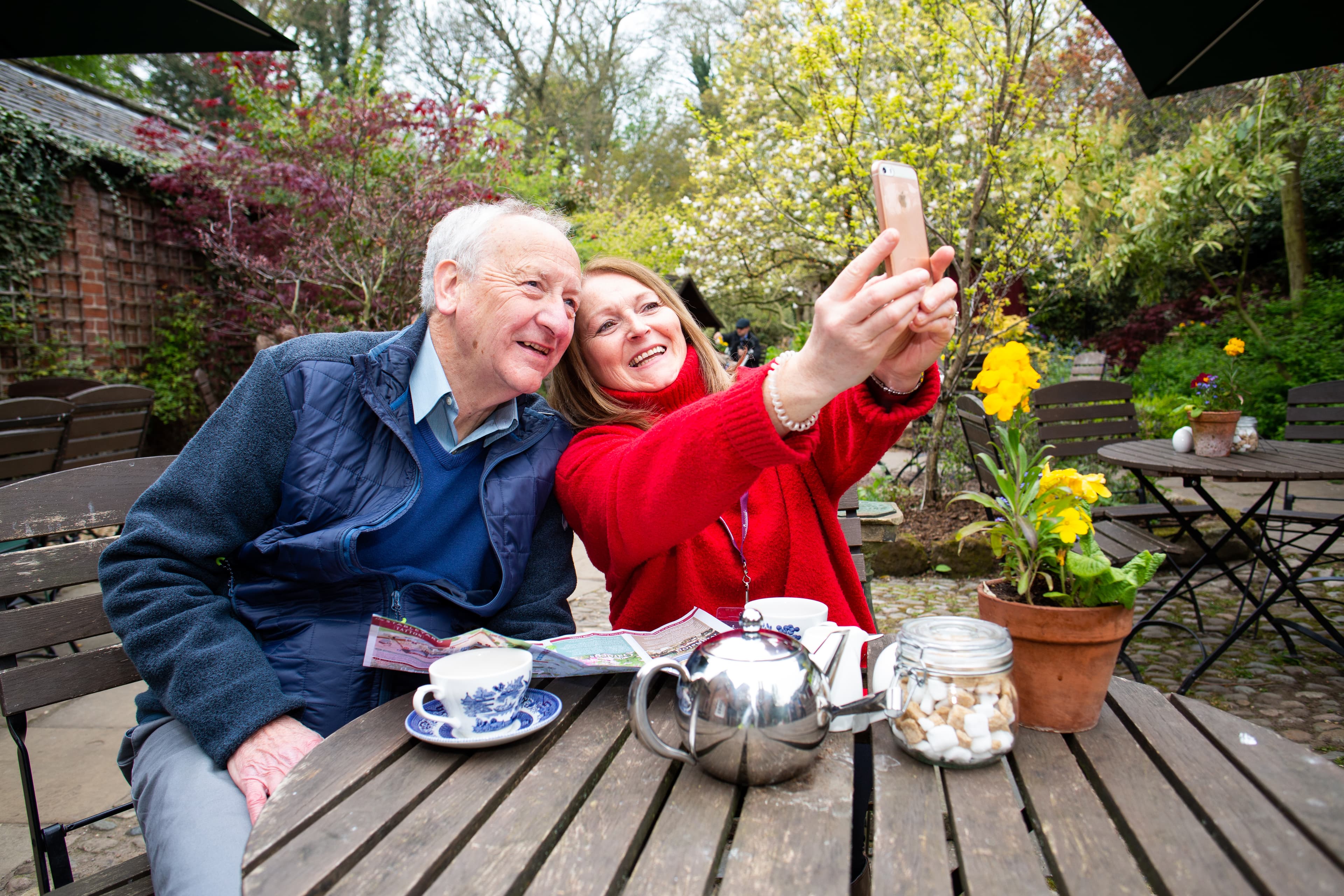 An elderly man and a woman take a selfie together at an outdoor café table with teacups and a teapot. - Home Instead