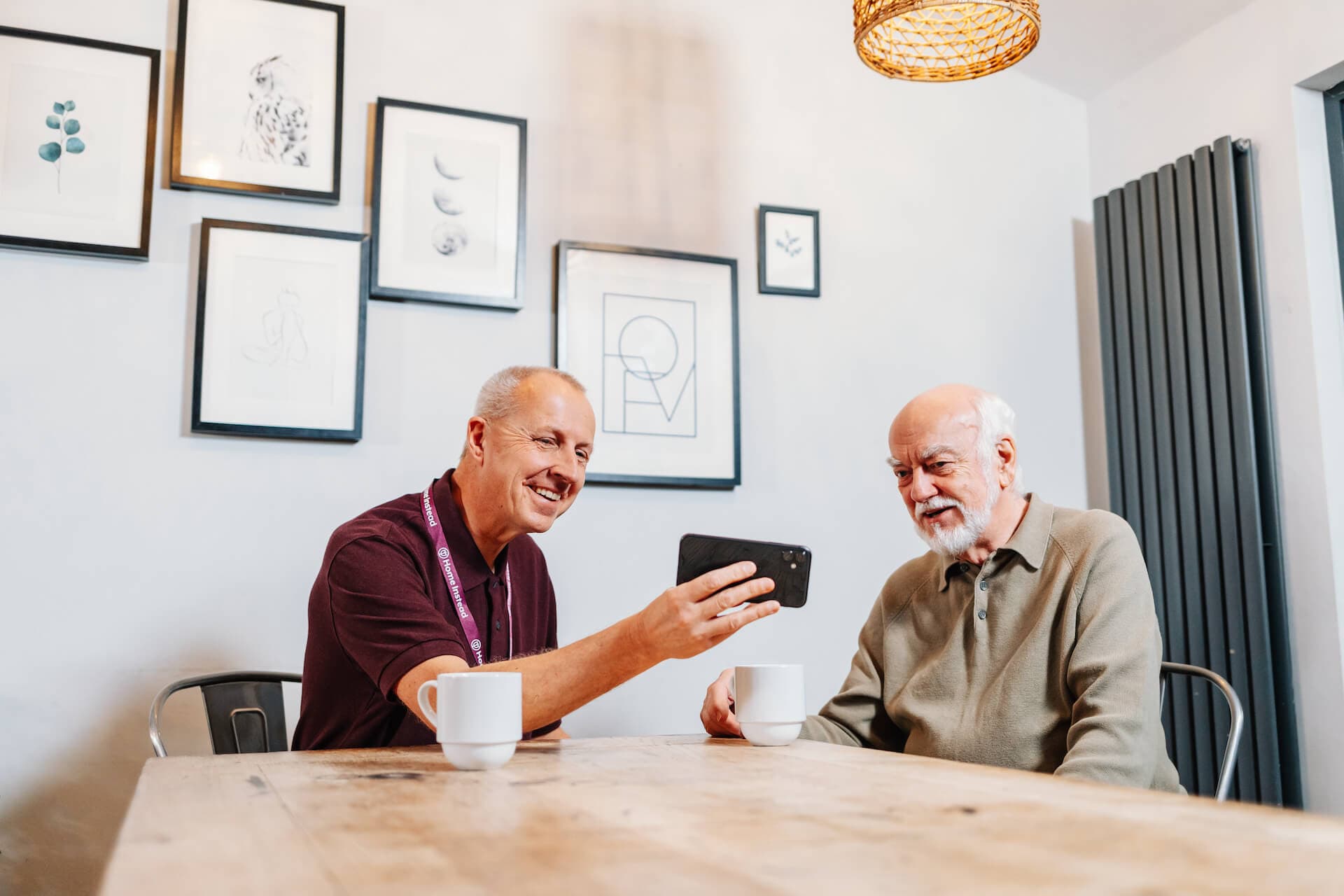 Two men sitting at a table, one showing a phone to the other with coffee cups in front of them and framed art on the wall. - Home Instead
