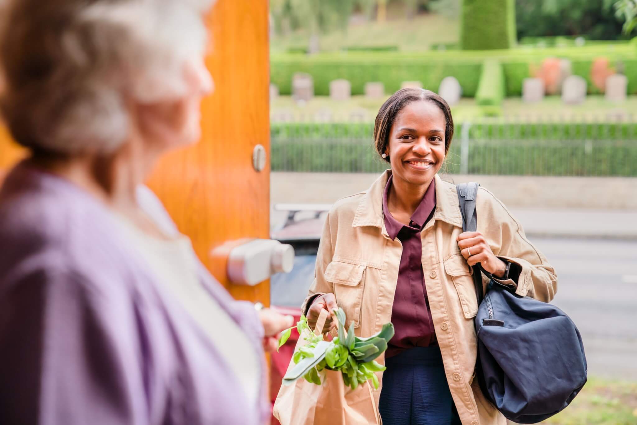 A woman smiles as she carries groceries and a bag, standing at an open door where an elderly woman stands. - Home Instead