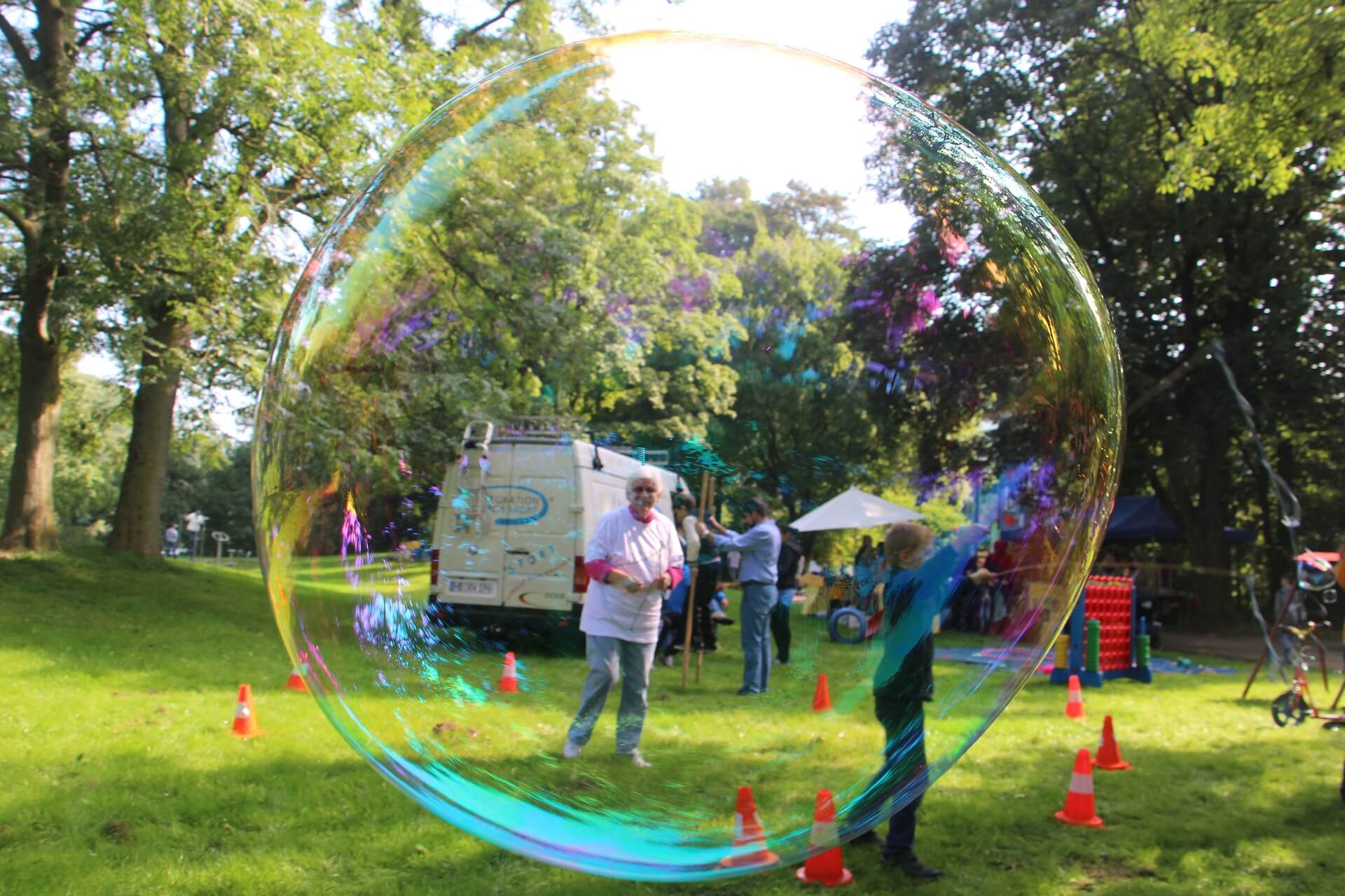 A person stands inside a giant bubble in a park, with trees and an ice cream van in the background. - Home Instead