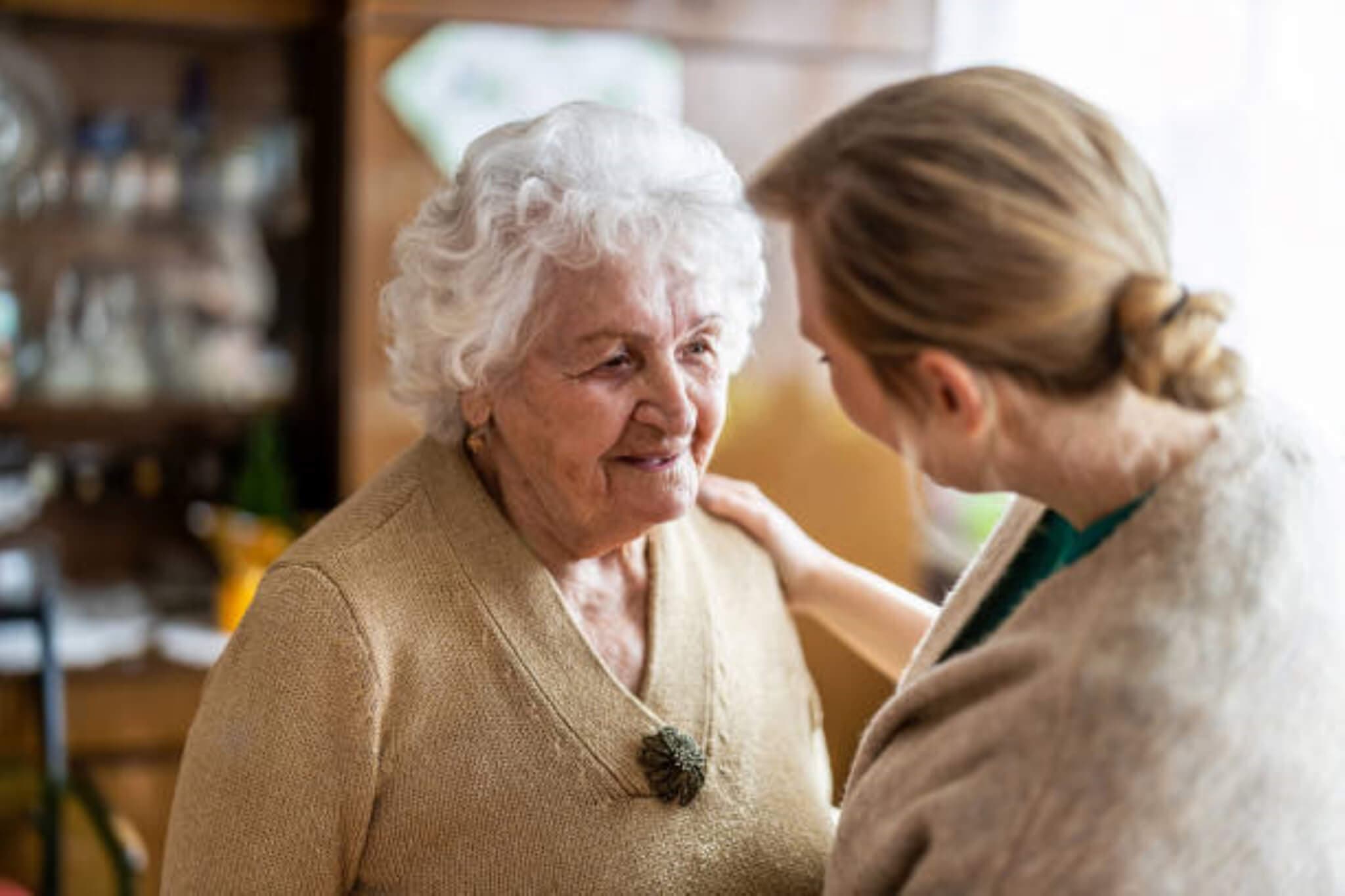 Elderly woman with white hair being comforted by another woman in a cozy indoor setting. - Home Instead