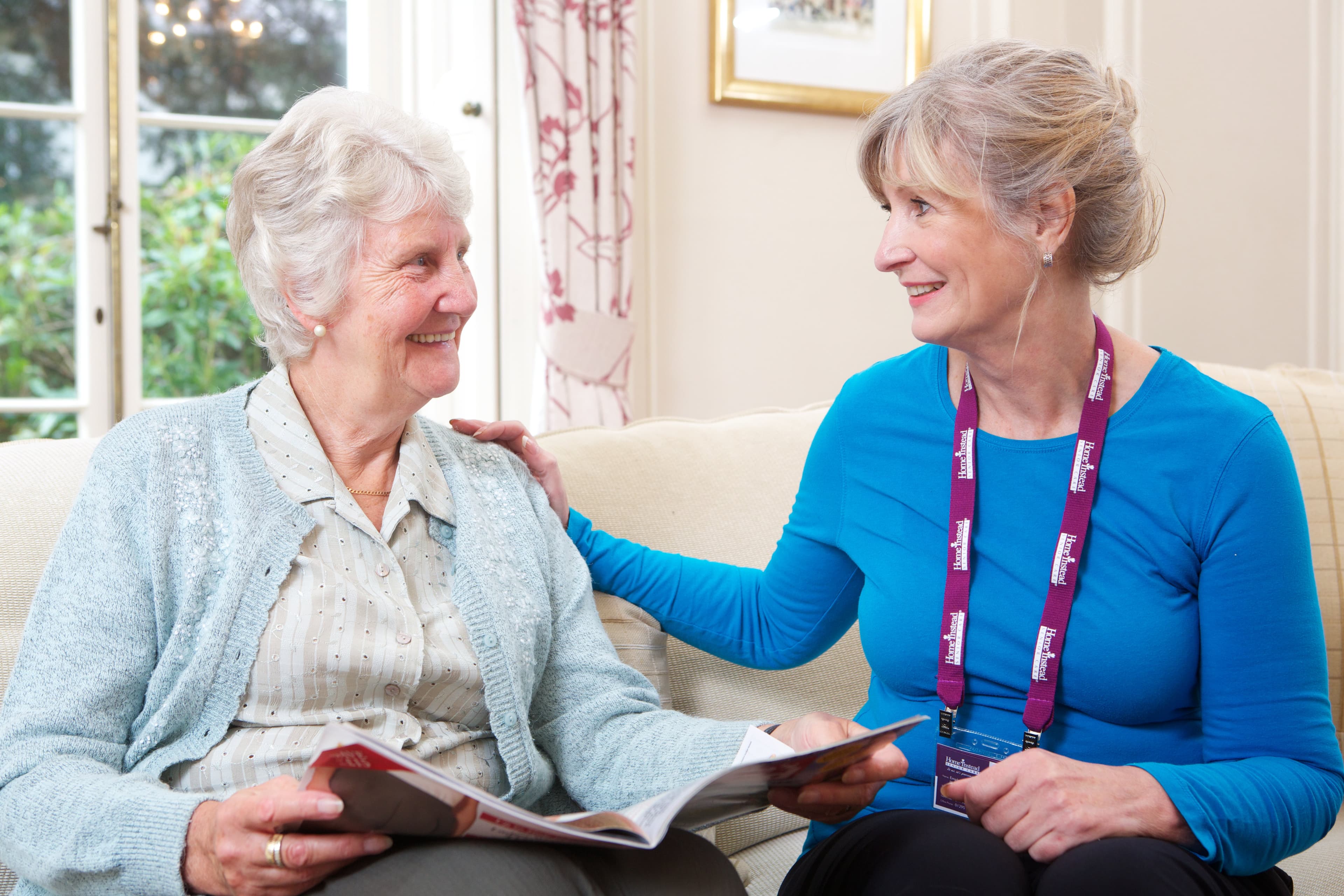 An elderly woman holding a magazine smiles at a caregiver who is sitting beside her, wearing a blue shirt and a name badge. - Home Instead