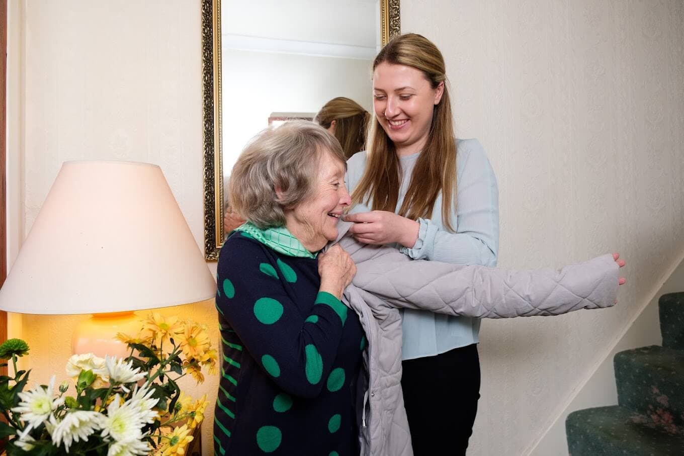 A younger woman helps an older woman put on a coat while they both smile in a warmly lit room. - Home Instead