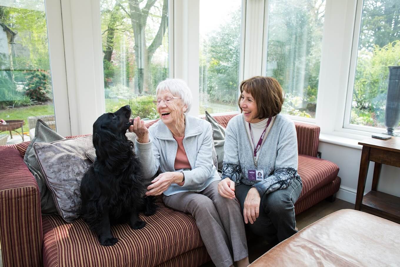 An elderly woman and a caregiver are sitting on a couch with a black dog, smiling and enjoying time together. - Home Instead