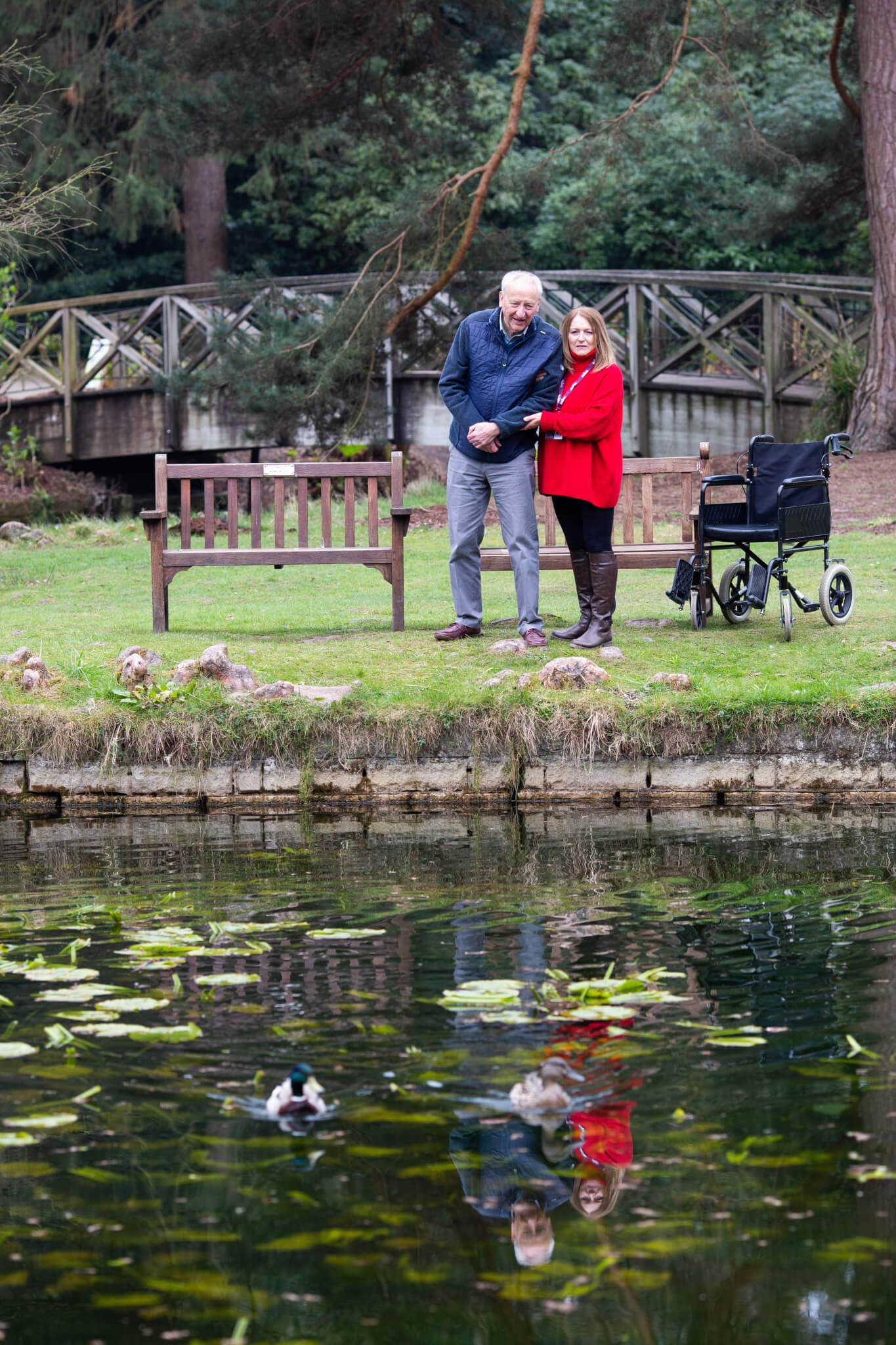 Two people stand by a pond feeding ducks. An empty wheelchair is nearby, and a wooden bridge is in the background. - Home Instead